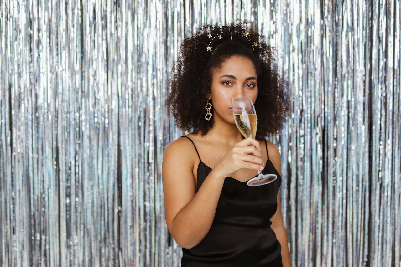 about-02 African American woman in a black dress celebrates with champagne against a silver backdrop.