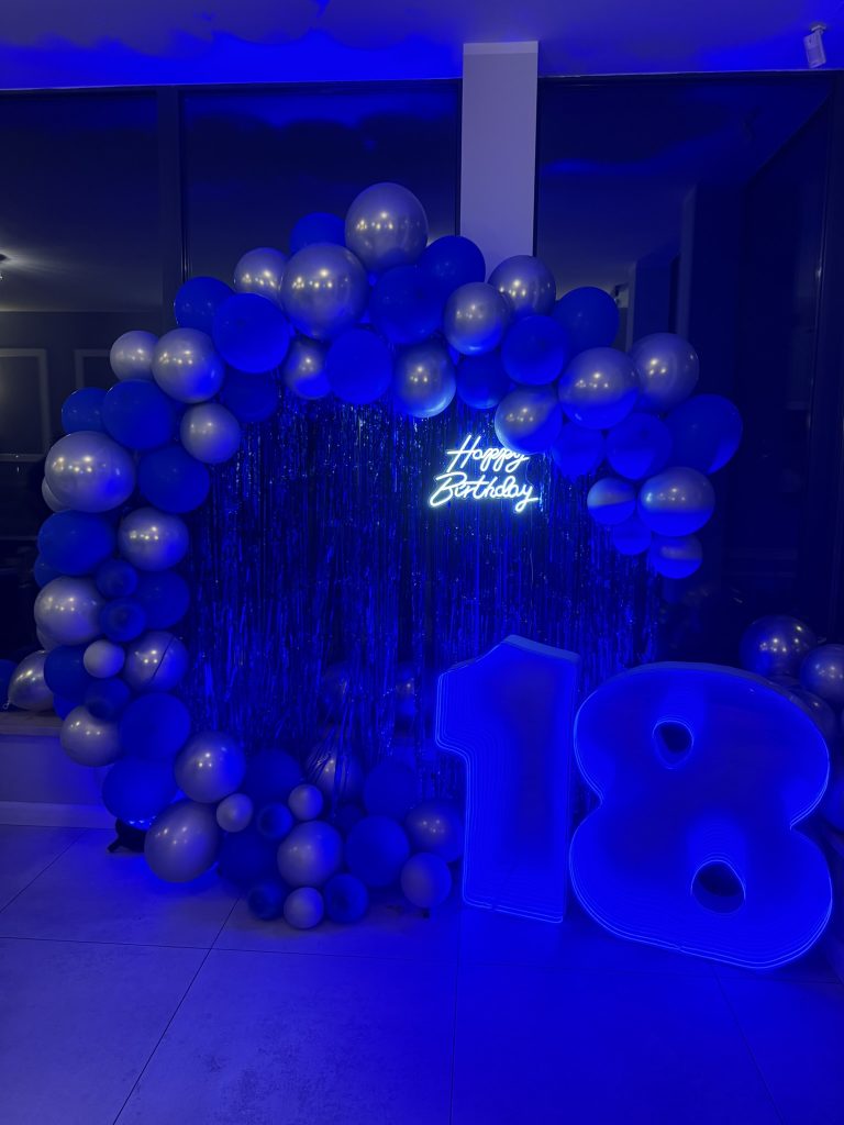 Świecąca cyfra LED 18 na tle niebieskiej ścianki balonowej – dekoracja na osiemnastkę latem
